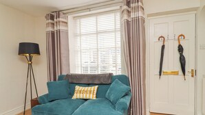 A teal sofa with a cushion, a floor lamp, and curtains in front of a door with two umbrellas hanging on hooks. - Birch Cottage (Skipton)