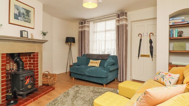 A cozy living room with a fireplace, a blue sofa, yellow ottoman, and a bookshelf. - Birch Cottage (Skipton)