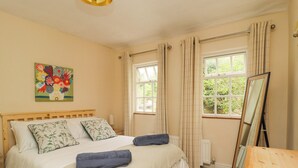A bedroom with a bed, two pillows, a blanket, a window with curtains, a mirror, and a painting on the wall. - Birch Cottage (Skipton)