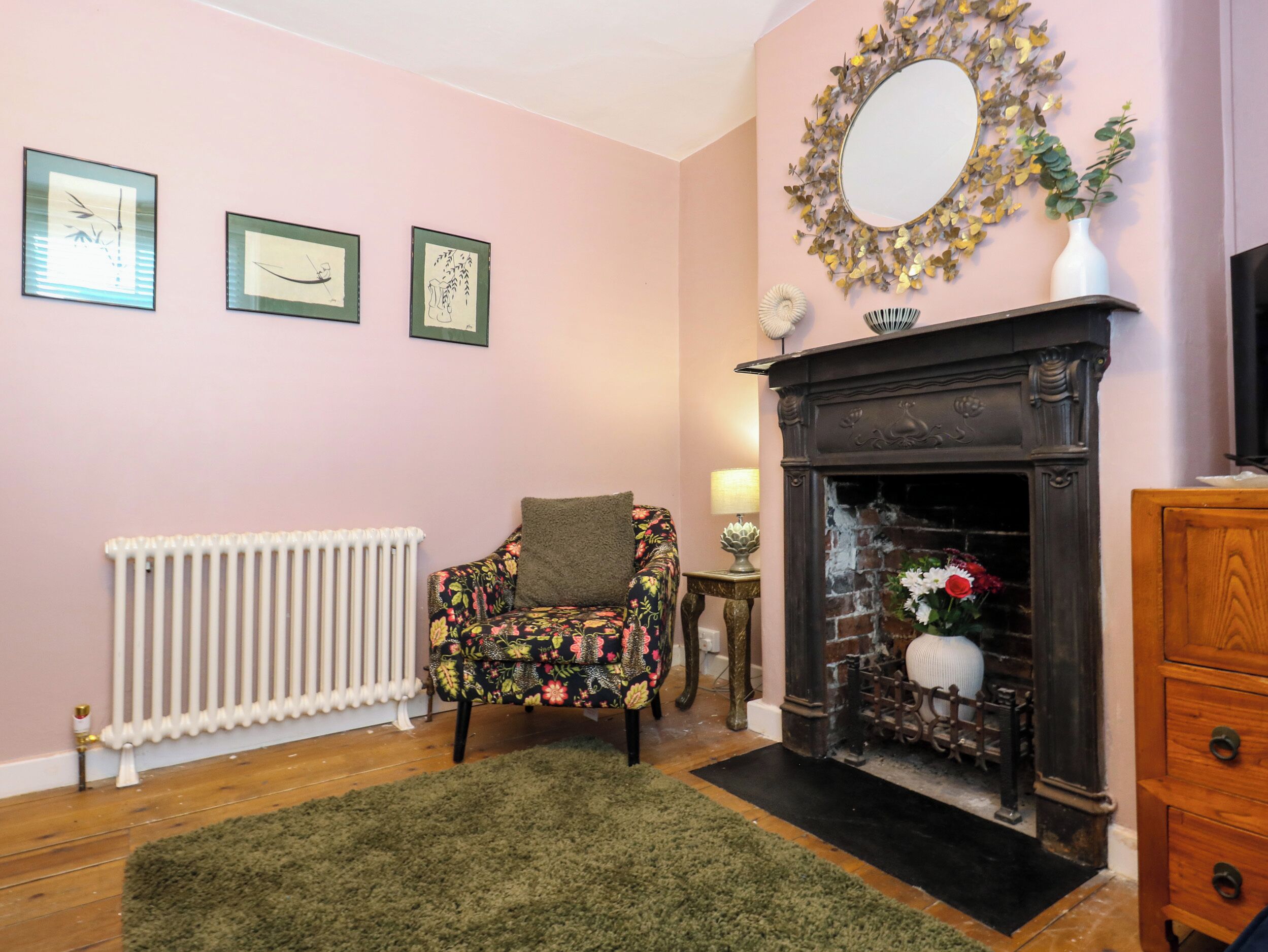 A cozy room with a fireplace, a radiator, a floral armchair, and three framed artworks on the wall.