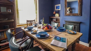 Uma sala de jantar com uma mesa de madeira posta para quatro pessoas, rodeada por cadeiras. Uma estante de livros e uma lareira são visíveis ao fundo.