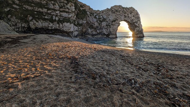 Paisagem costeira com um arco natural de rocha, uma praia de areia e o oceano.