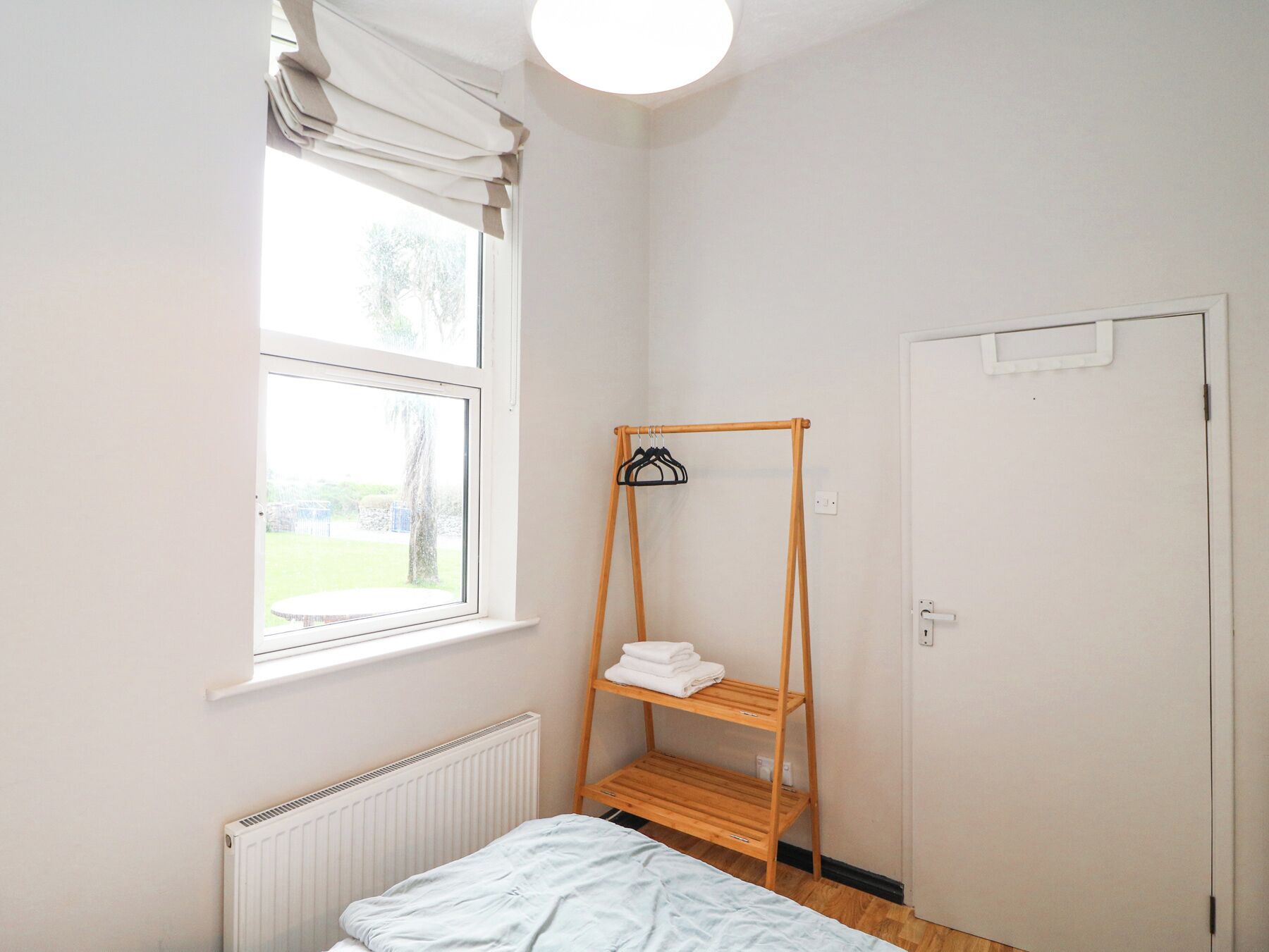 A small, bright room with a single bed, a window with curtains, a radiator, and a wooden shelf with towels.