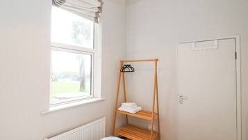A small, bright room with a single bed, a window with curtains, a radiator, and a wooden shelf with towels.