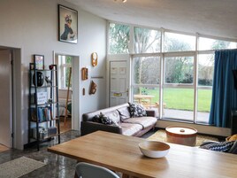 A spacious living room with a large window, a wooden dining table, a sofa, and a bookshelf.