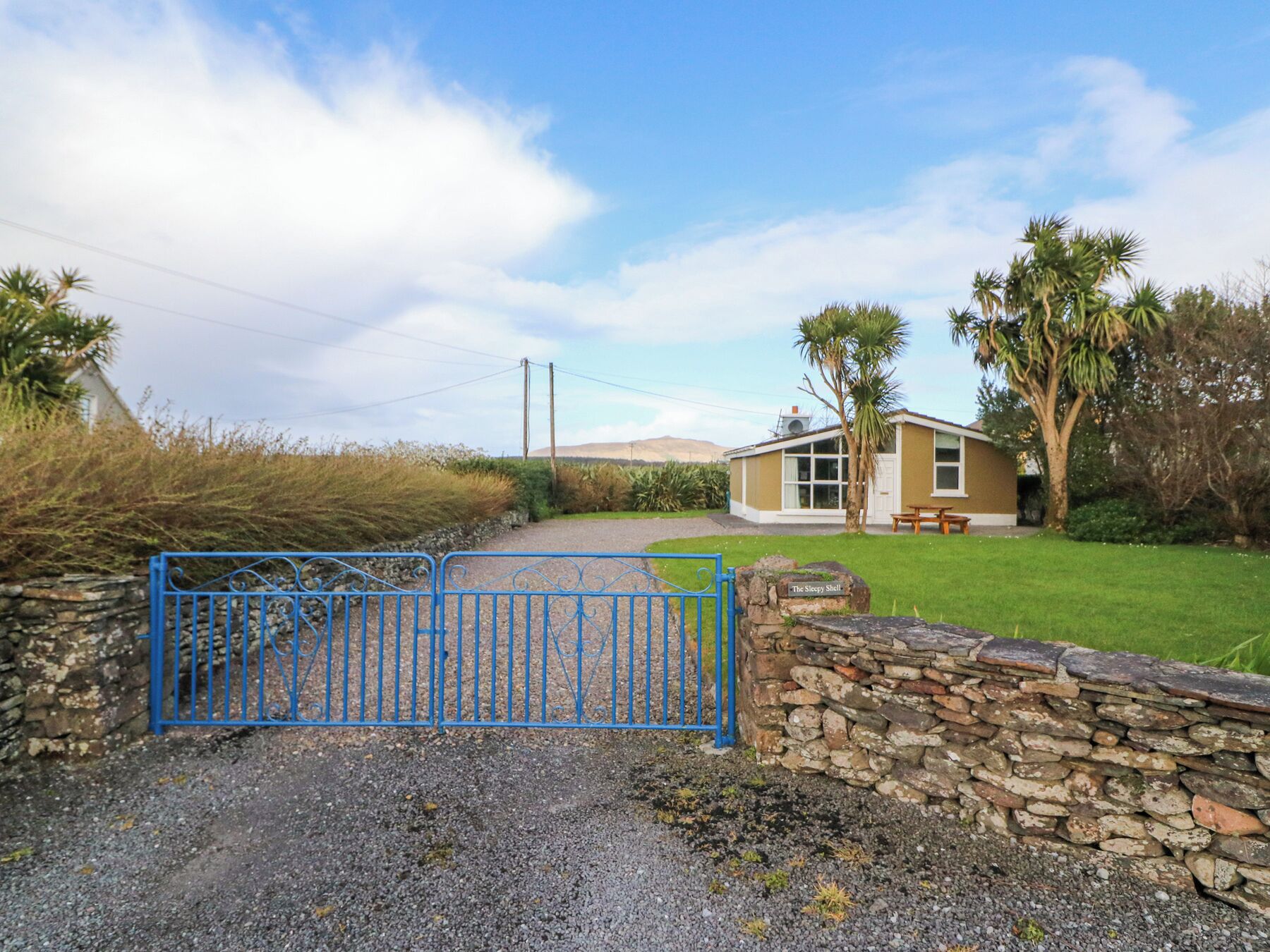 A rural property with a stone wall, a blue metal gate, and a small building with a bench and a table outside.