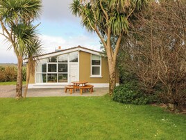 A small, single-story house with a conservatory, a patio with a wooden table and benches, and a lawn.