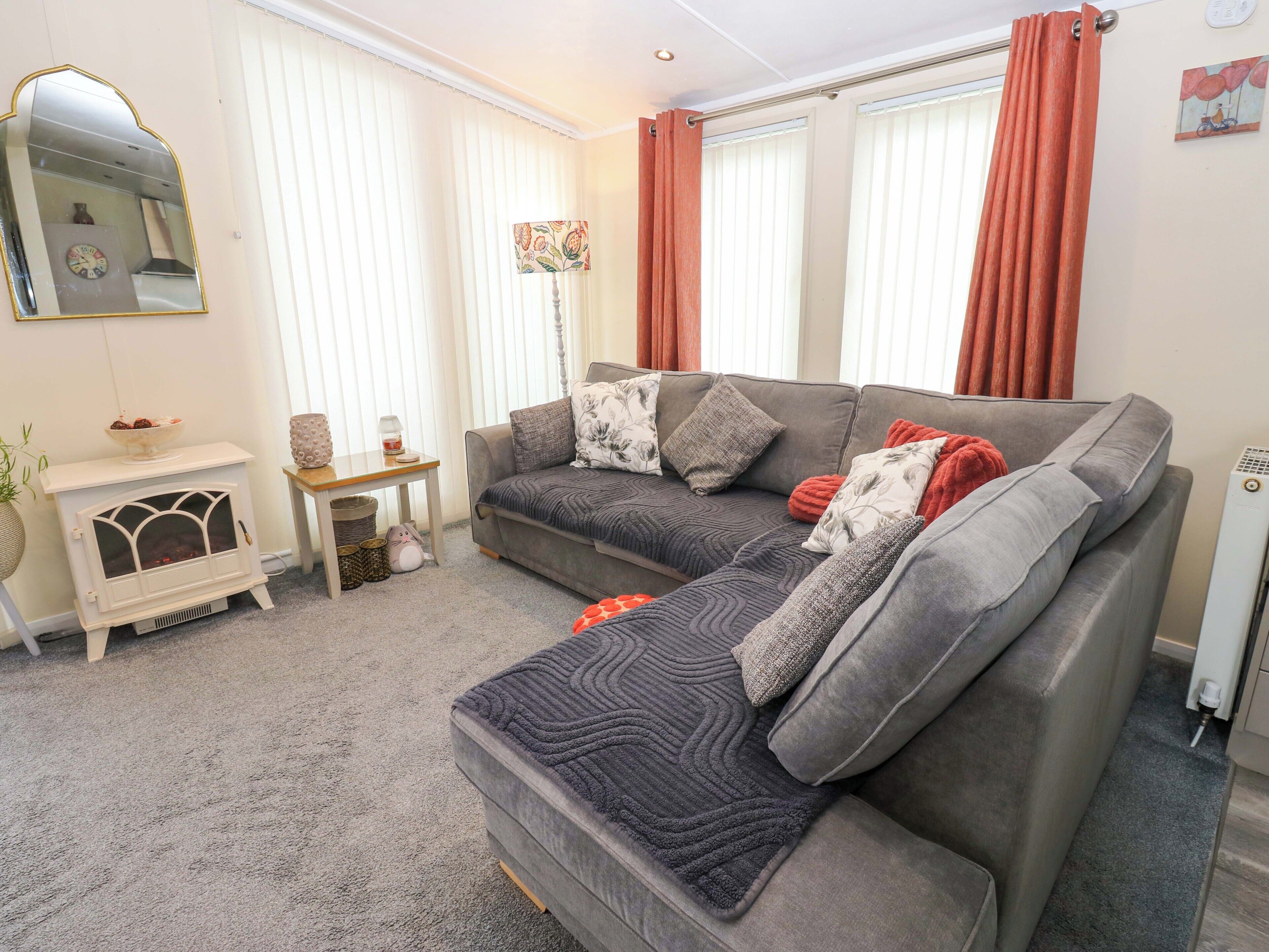 A living room with a grey sofa, a white fireplace, a small table with a lamp, and a wall-mounted mirror.