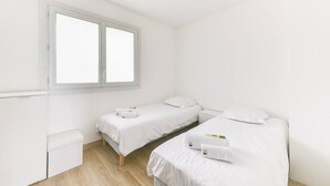 A clean, white room with two single beds, a small bedside table, and a window.