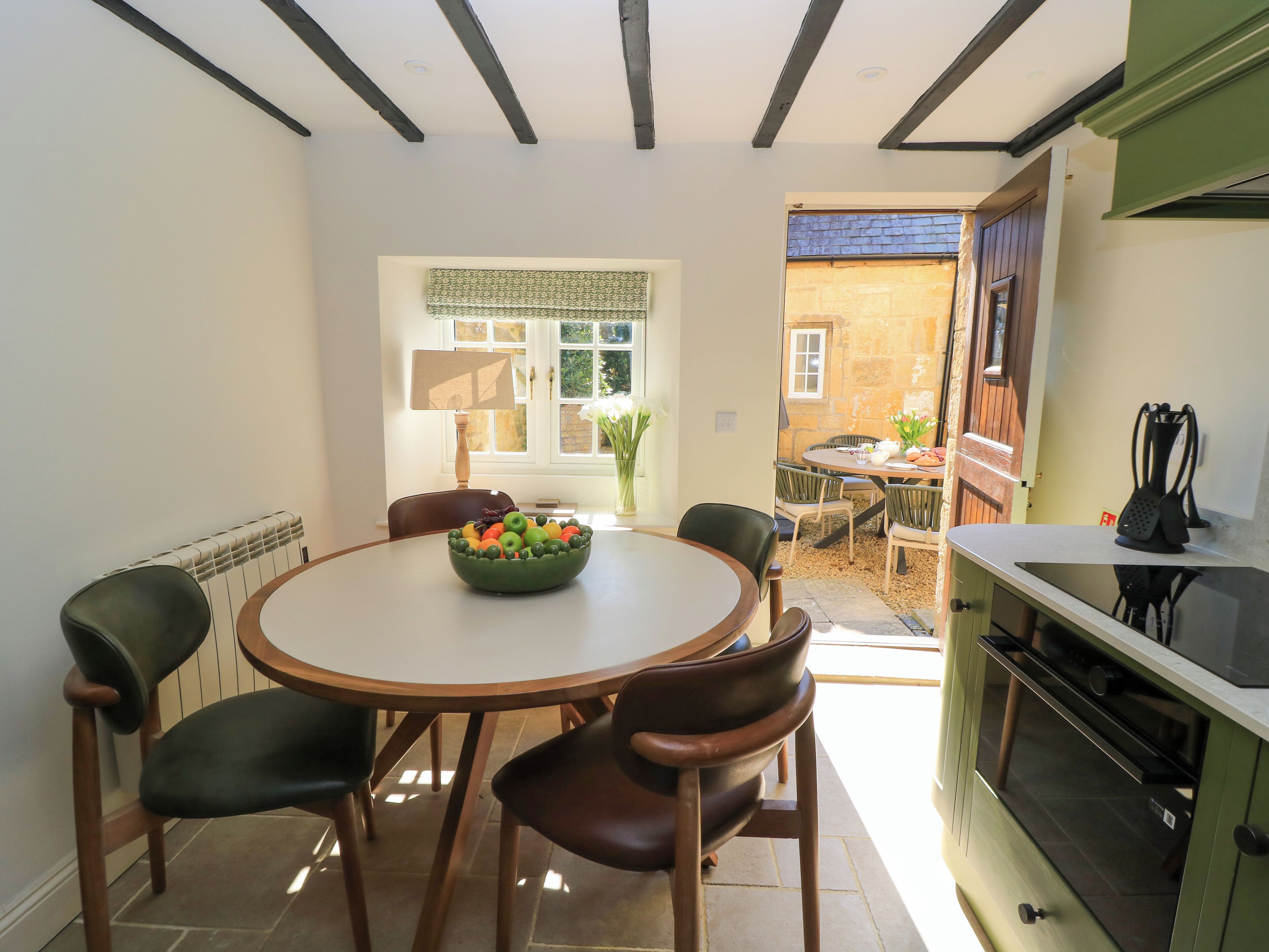 A dining area with a round table, chairs, and a bowl of fruit. There is a window with a view of a garden and a door leading to an outdoor area.