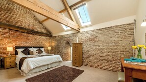 A bedroom with a brick wall, wooden beams, a bed with pillows, a nightstand, a dresser, a desk with a lamp, and a window. - Coot (Beccles)