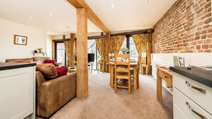 A cozy living area with a brown leather sofa, wooden dining table, and chairs. A kitchen area with white cabinets and a brick wall is visible. - Woodcock (Beccles)