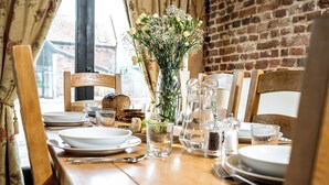 A dining table set with plates, glasses, and a vase of flowers, with a brick wall and window in the background. - Woodcock (Beccles)