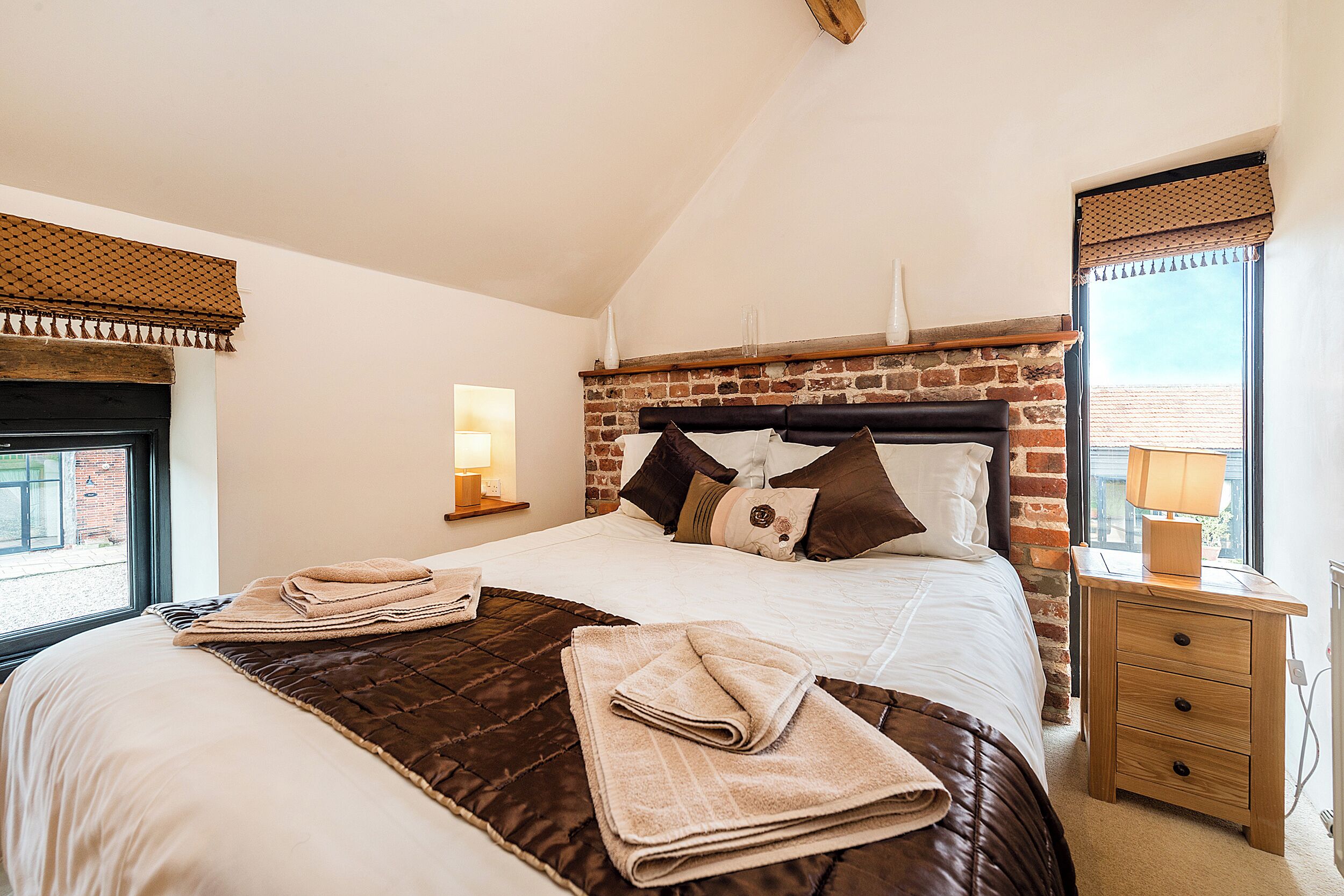 Un lit bien fait, recouvert d’une couverture marron, une table de chevet en bois avec une lampe, et un mur de briques derrière le lit.