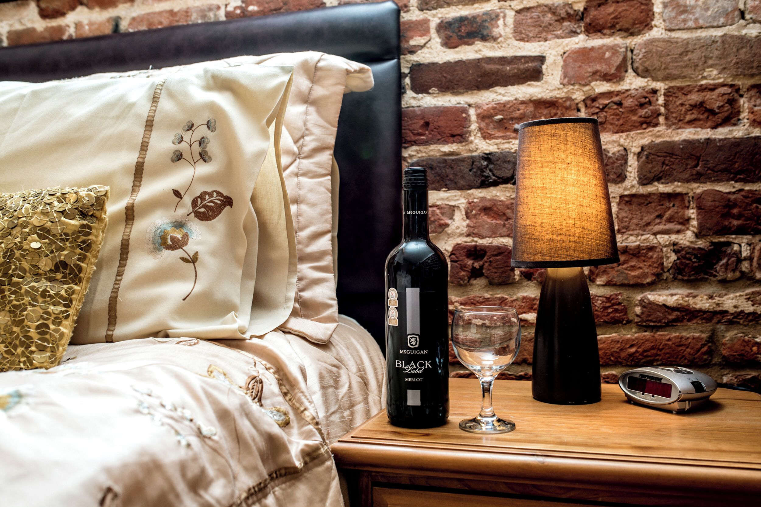 Una cama bien hecha con una almohada decorativa, una botella de vino, una copa de vino y una lámpara sobre una mesita de noche de madera frente a una pared de ladrillo.