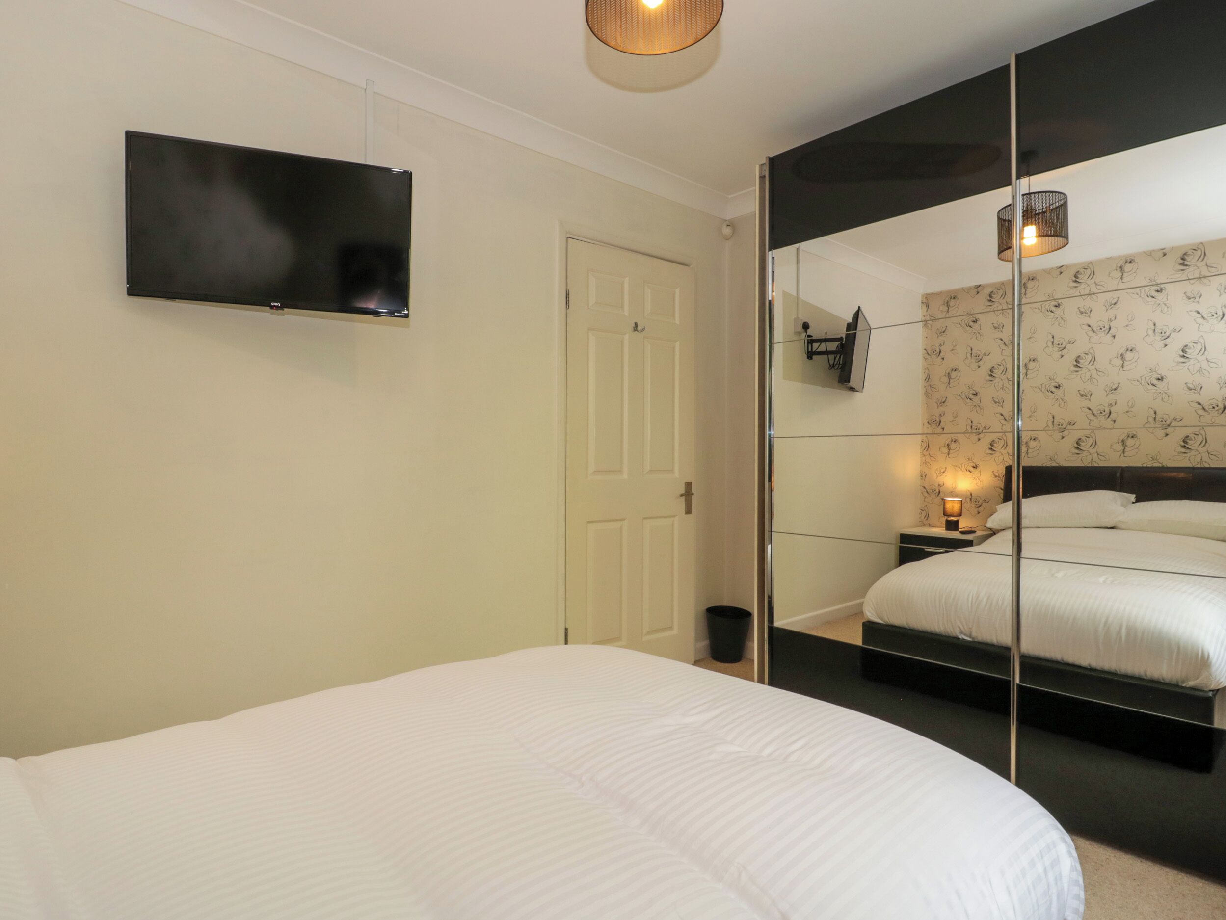 A bedroom with a bed, a TV mounted on the wall, a mirror reflecting the room, and a nightstand with a lamp.