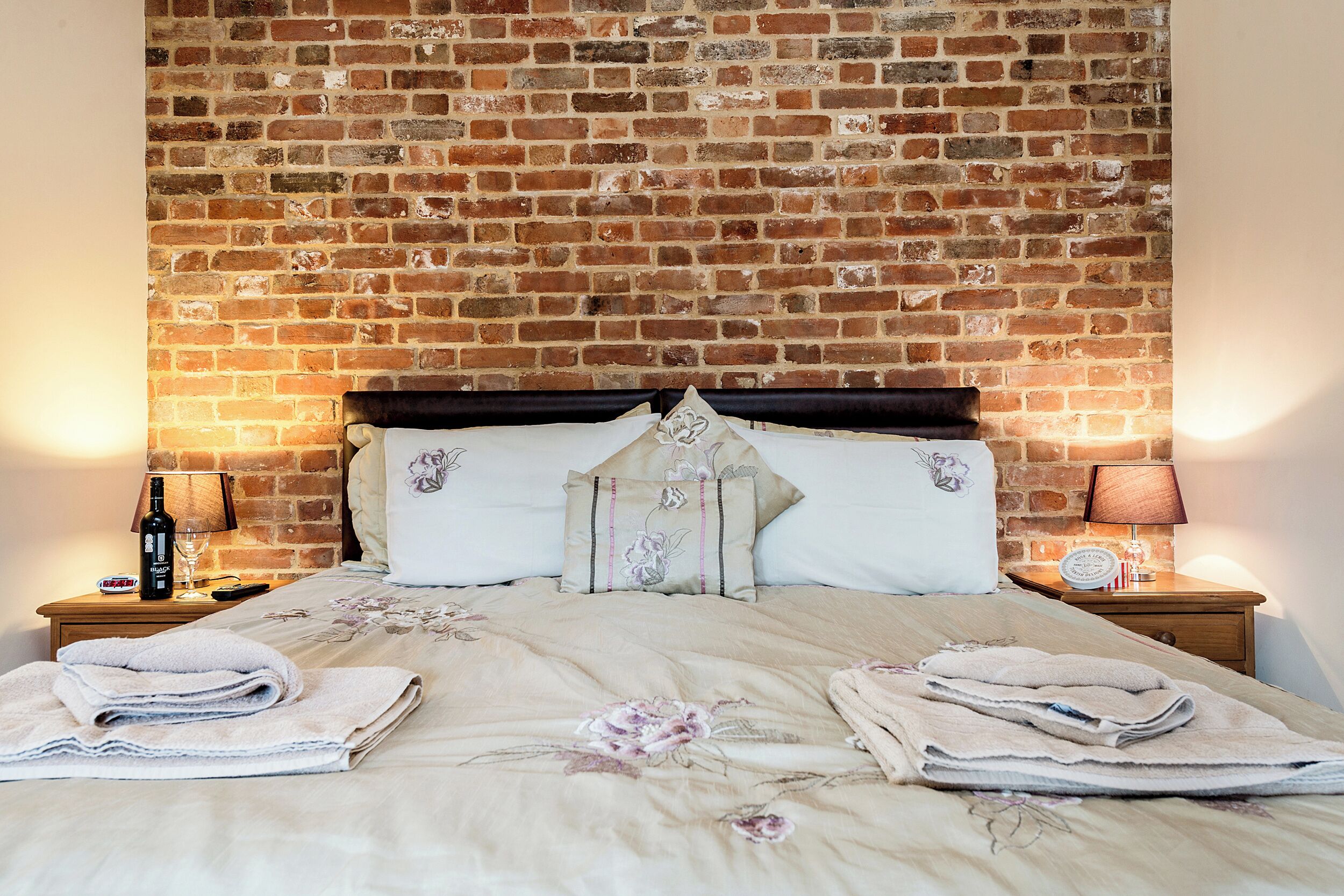 A neatly made bed with floral bedding, a brick wall behind, and two bedside tables with lamps and a bottle.
