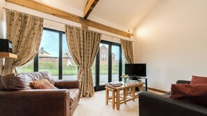 A living room with a brown leather sofa, a wooden coffee table, and a black leather couch. There is a TV on a stand and a window with curtains. - Partridge (Beccles)