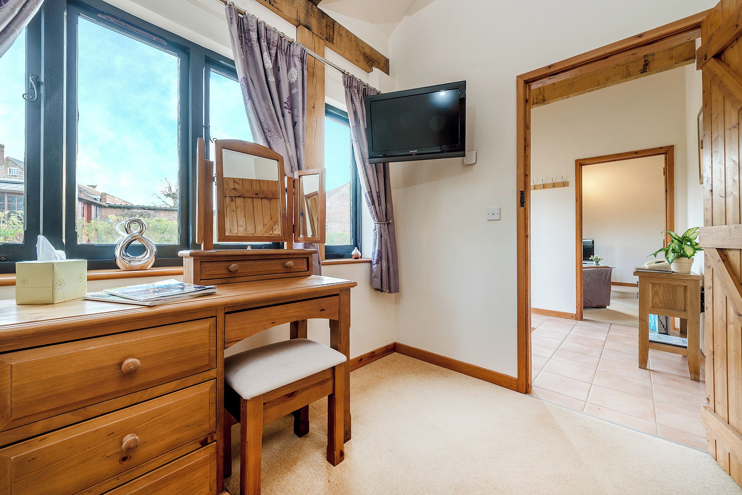 A room with a wooden desk, a television mounted on the wall, and a large window with curtains.
