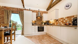 A kitchen with white cabinets, a brick wall, a wooden dining table, and a view of greenery outside. - Partridge (Beccles)