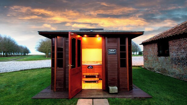 A wooden sauna structure with a bench inside, surrounded by a grassy area and a brick building in the background. - Partridge (Beccles)