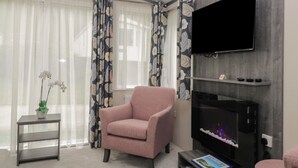 A modern living room with a pink armchair, a flat-screen TV mounted on a grey wall, a small side table with a vase of flowers, and a bookshelf with decorative items. - Caravan 7 (Plymouth)