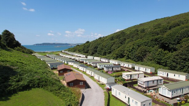 A caravan park with rows of caravans, a pathway, and a view of the sea and hills. - Caravan 7 (Plymouth)