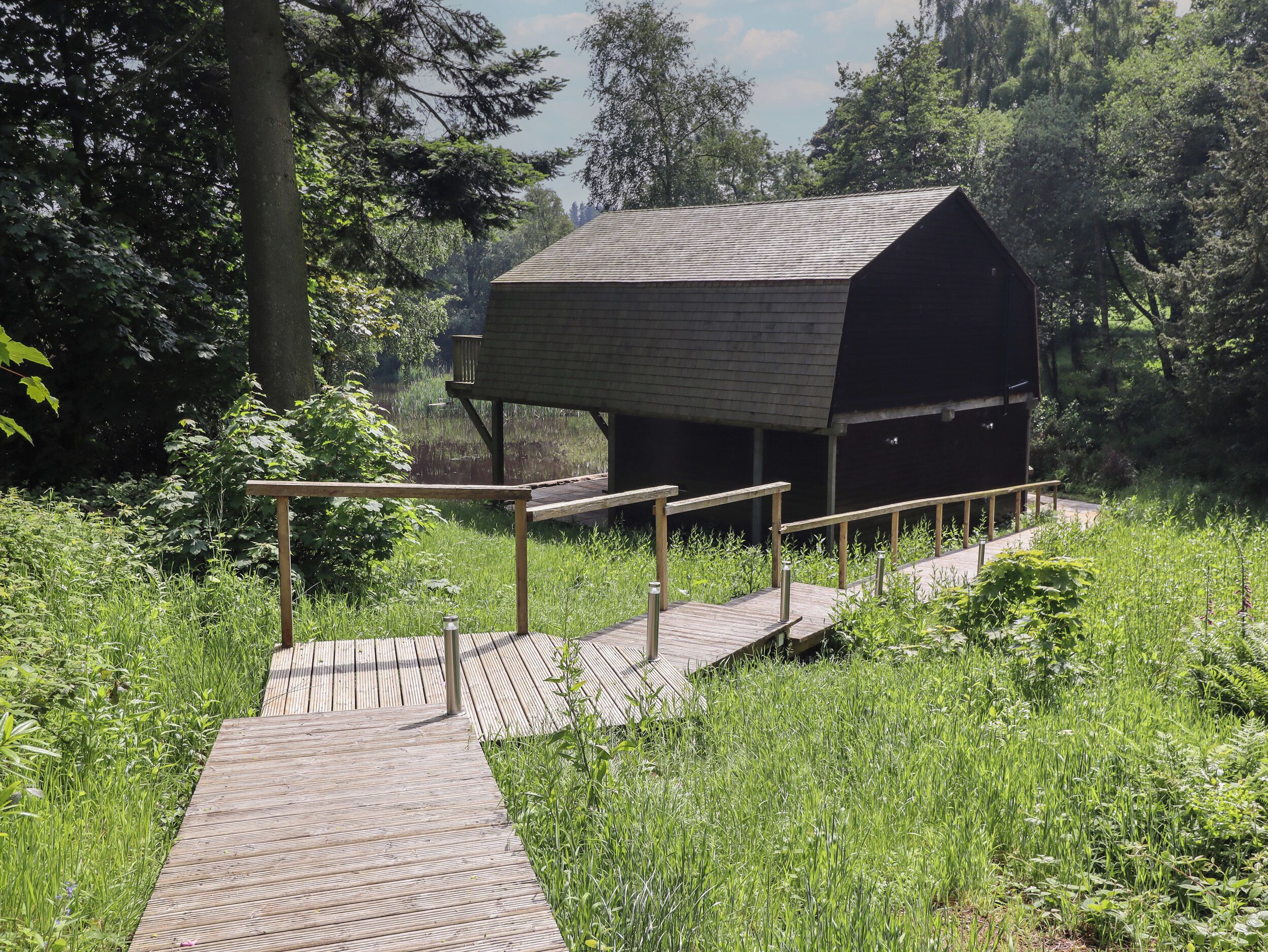 A wooden deck leading to a covered shelter with a sloped roof, surrounded by lush greenery and trees.