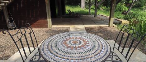 Una mesa redonda exterior con cubierta de mosaico, rodeada de sillas de metal, bajo una pérgola de madera.