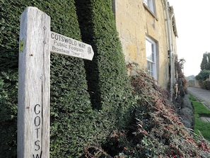 A wooden signpost with 'COTSWOLD WAY' and 'Broadway Tower 1½' written on it, set against a backdrop of a building and dense foliage. - Bay House Cottage (Broadway)