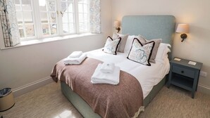 A neatly made bed with pillows, a bedside lamp, a nightstand, and a window with curtains. - Bay House Cottage (Broadway)