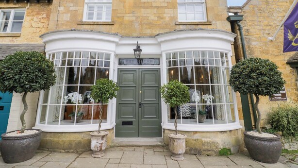 A corner building with a green door, two large windows with white frames, and potted plants. - Bay House Cottage (Broadway)