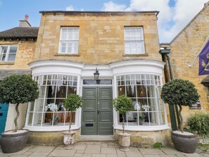 A corner building with a green door, two large windows with white frames, and potted plants. - Bay House Cottage (Broadway)