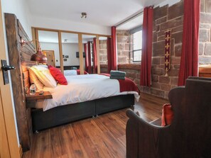 A bedroom with a large bed, wooden flooring, a stone wall, and red curtains. - Monastery Hall (Chester)