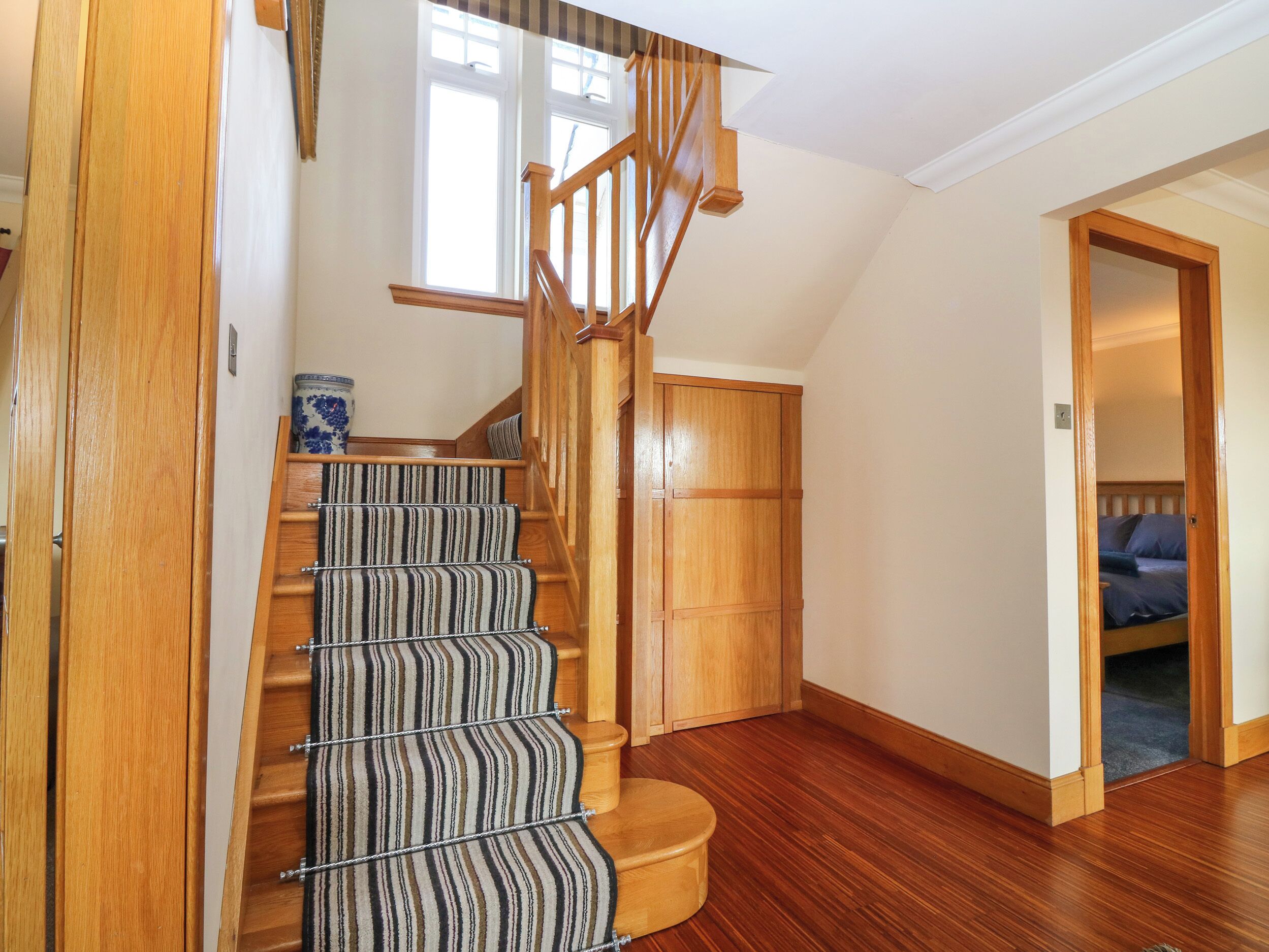 Una escalera de madera con alfombra a rayas que lleva a un dormitorio con puerta de madera y vista al cielo azul.