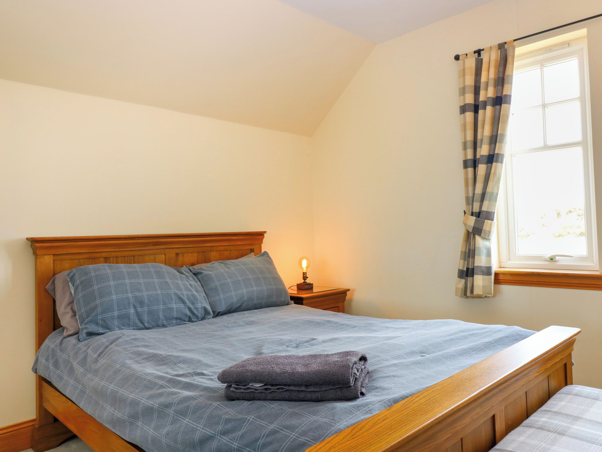 A neatly made bed with a blue checkered bedspread and pillows, a bedside lamp, and a window with curtains.