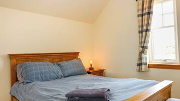 A neatly made bed with a blue checkered bedspread and pillows, a bedside lamp, and a window with curtains.