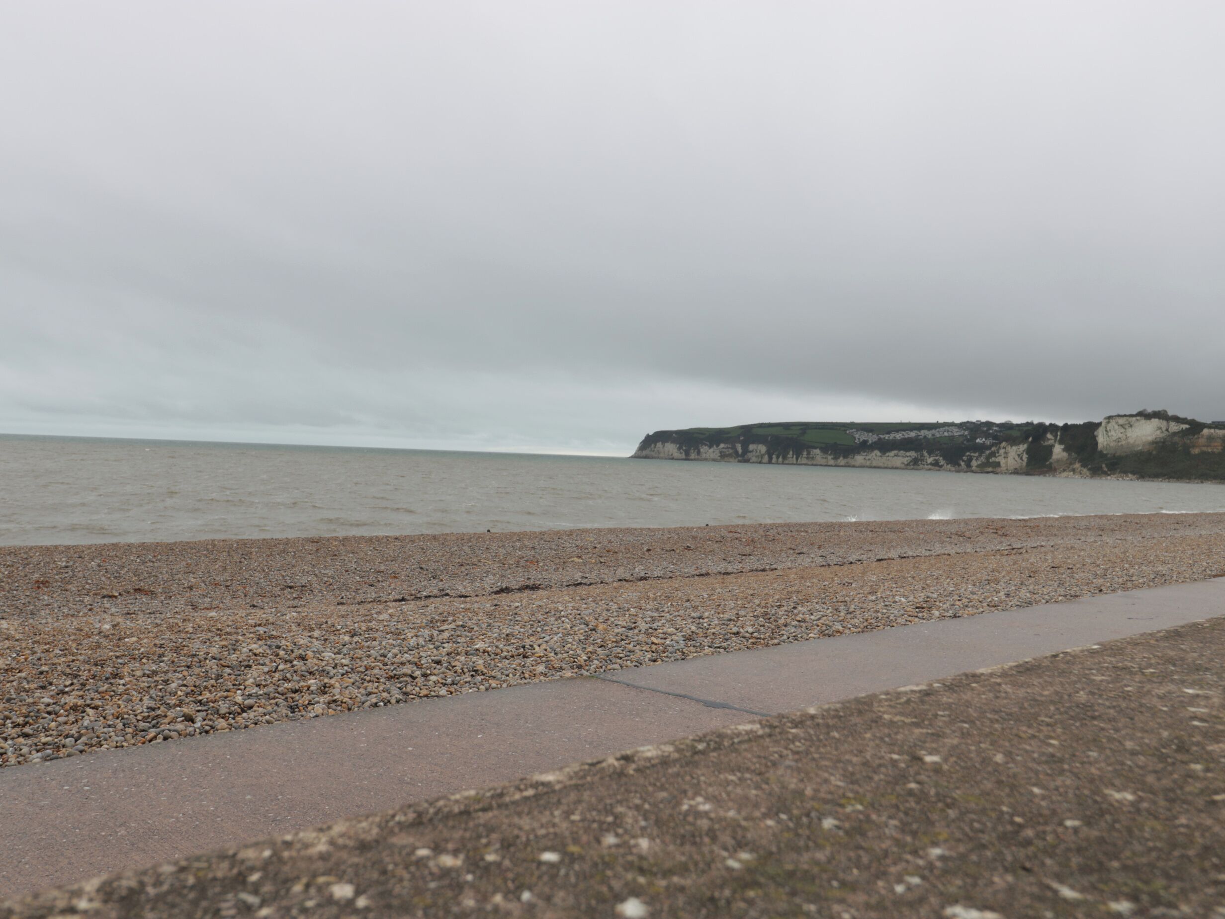 Une plage de galets avec un chemin de béton menant à l’eau, et des falaises en arrière-plan.