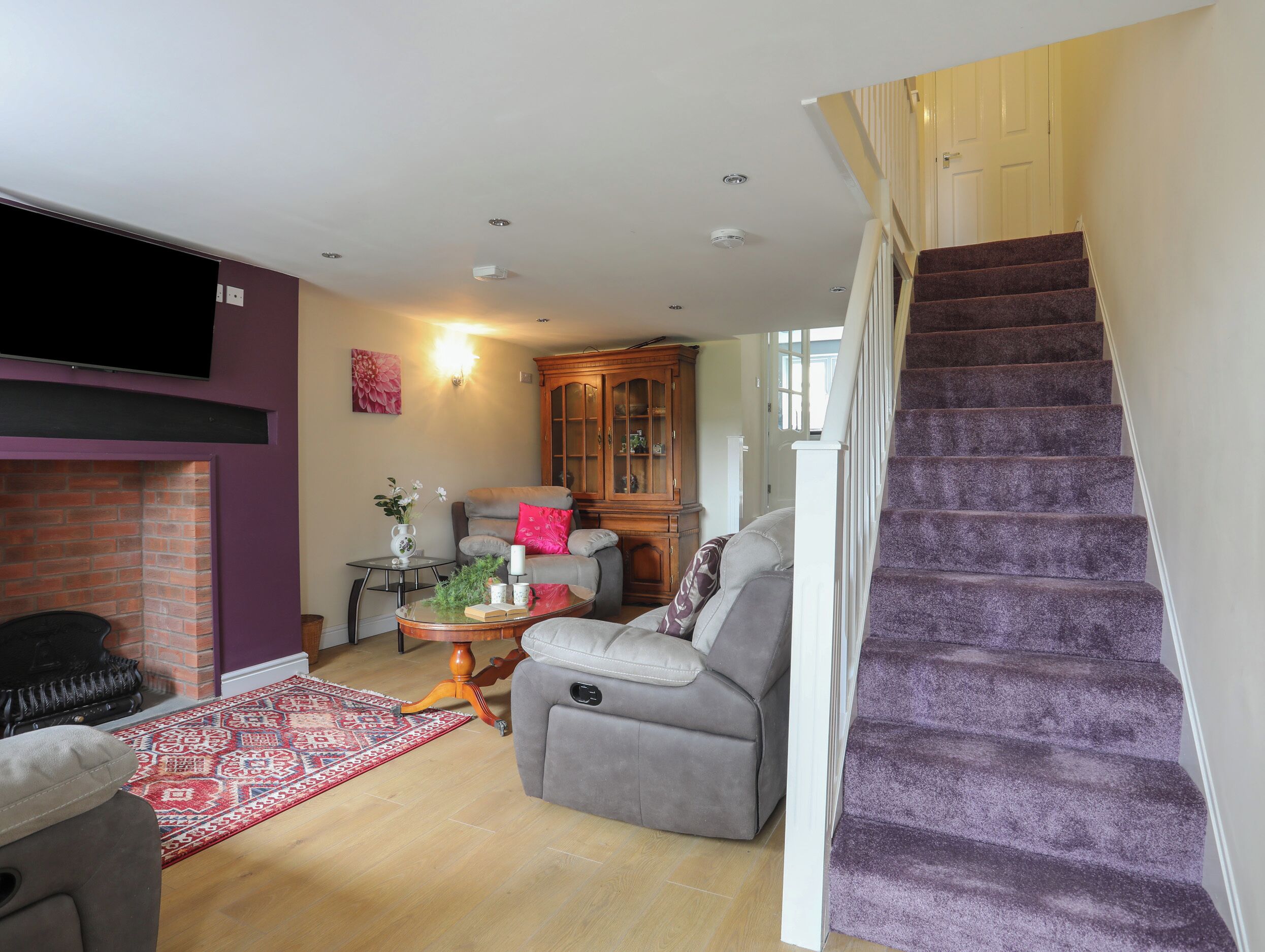 Un salon avec une cheminée, un canapé gris, un tapis rouge à motifs et un escalier recouvert d’une moquette violette.