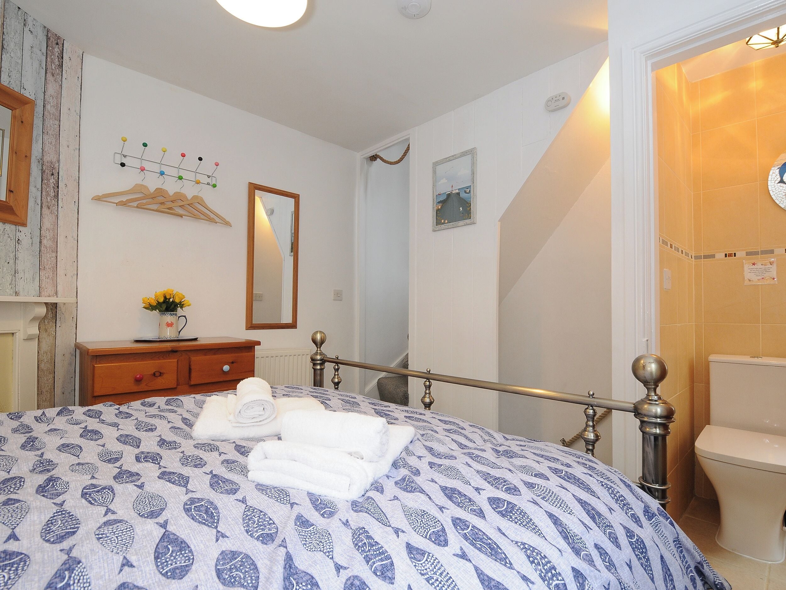 Ein Schlafzimmer mit Bett, Kommode, Spiegel und einem durchgehenden Türrahmen sichtbaren Badezimmer.