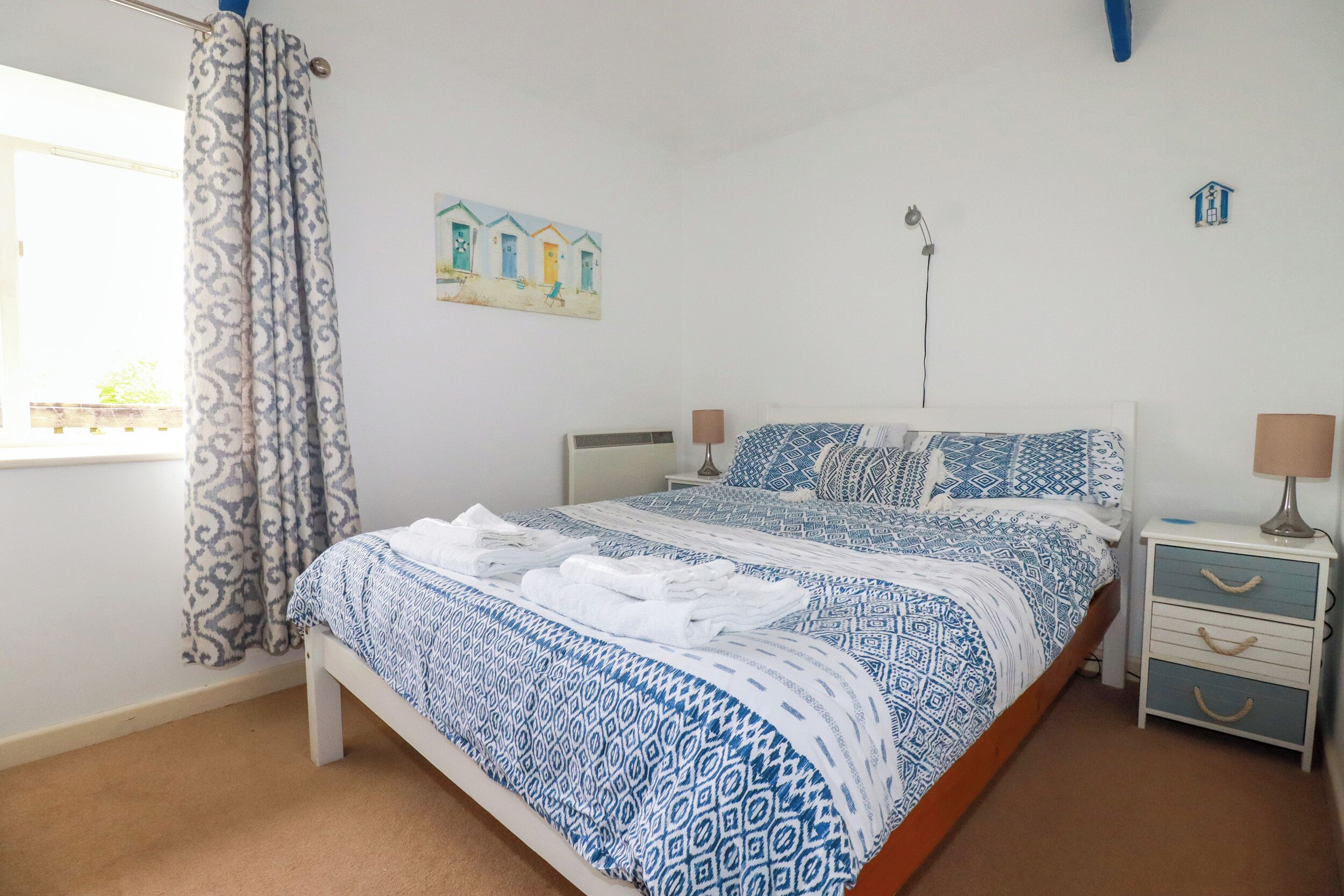 A bedroom with a bed, a nightstand, a lamp, a window with curtains, and a wall painting.