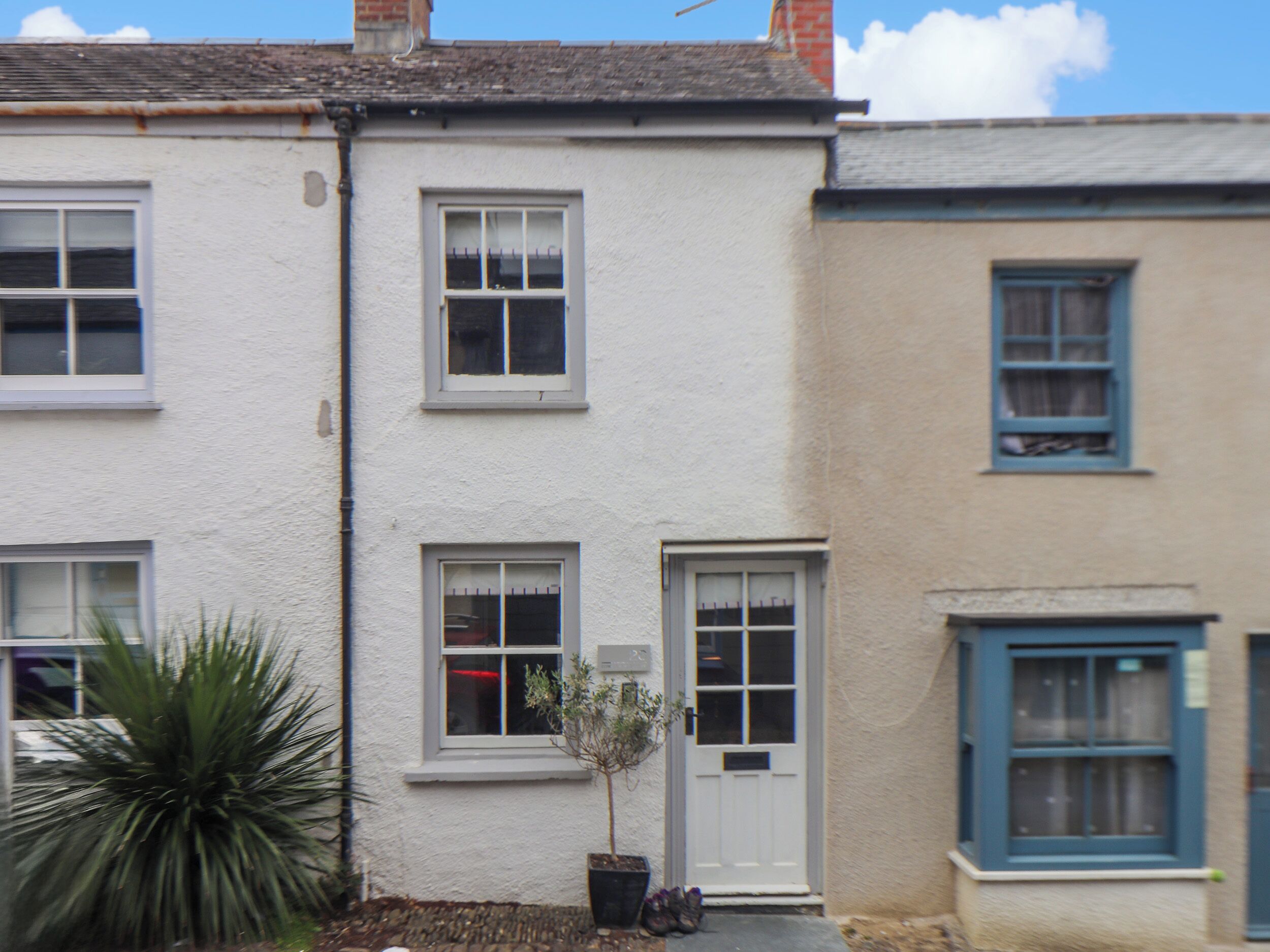 Une rangée de maisons mitoyennes aux façades blanches et beiges, dotées de nombreuses fenêtres et agrémentées d’un petit arbre dans le jardin avant.