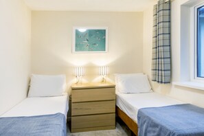 A bedroom with two single beds, a nightstand with two lamps, a wall-mounted picture, and a window with a curtain. - Chalet 7 at Landscove Holiday Park (Brixham)