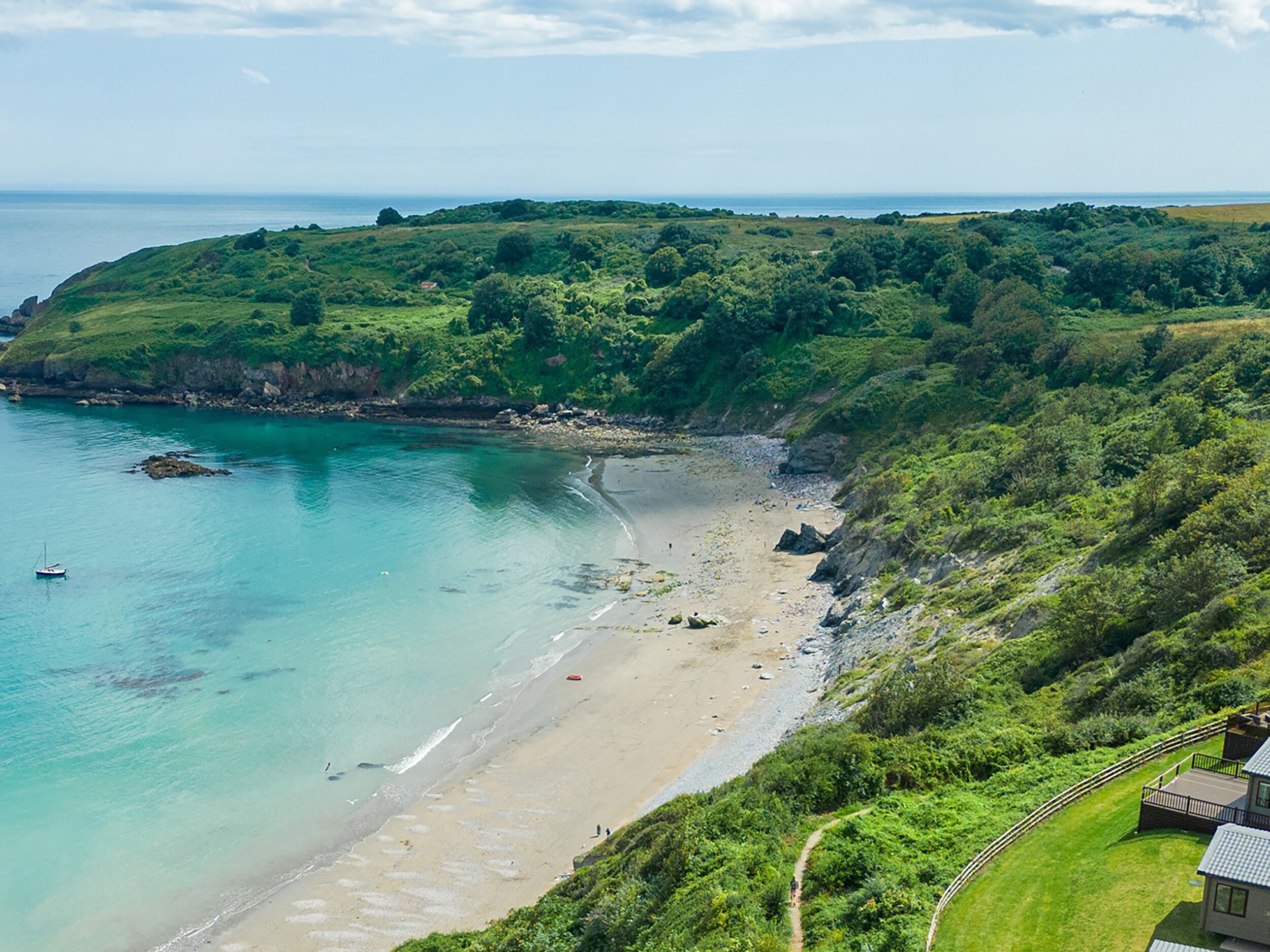 Un paisaje costero con una playa, vegetación y un edificio pequeño al que conduce un camino.