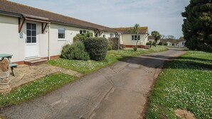 A row of single-story houses with a paved driveway, surrounded by greenery and white flowers. - Chalet 34 at Landscove Holiday Park (Brixham)