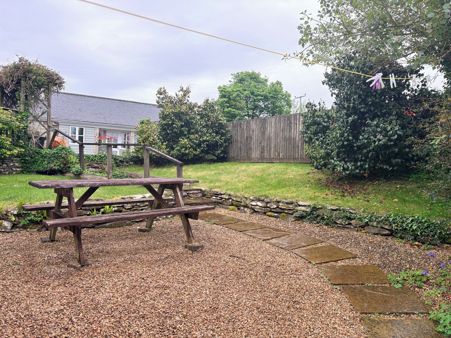 A picnic table in a garden with a stone border, a wooden fence, and a house in the background.