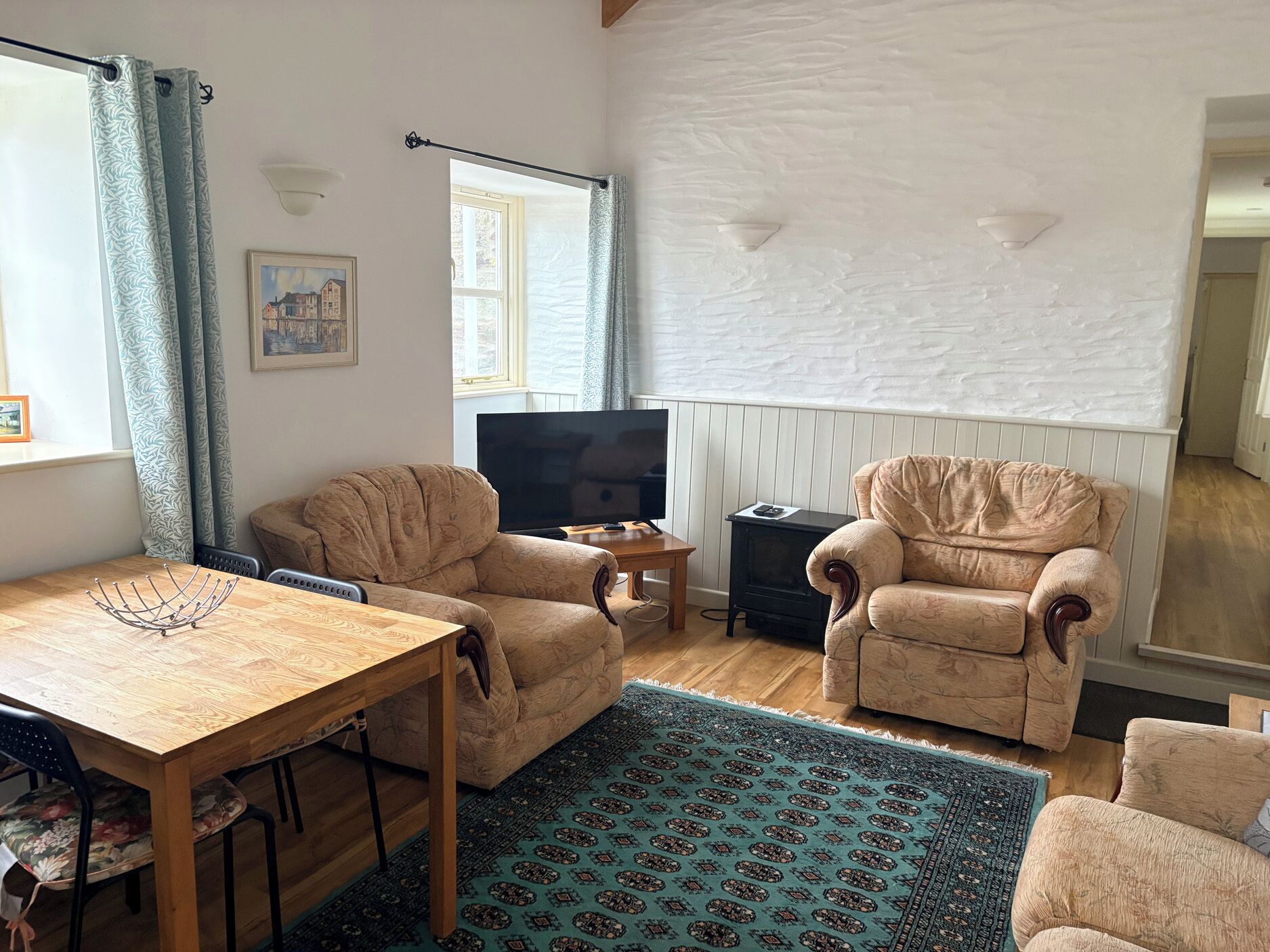 Uma sala de estar com uma mesa de madeira, duas poltronas, uma televisão, uma mesinha e um tapete estampado.