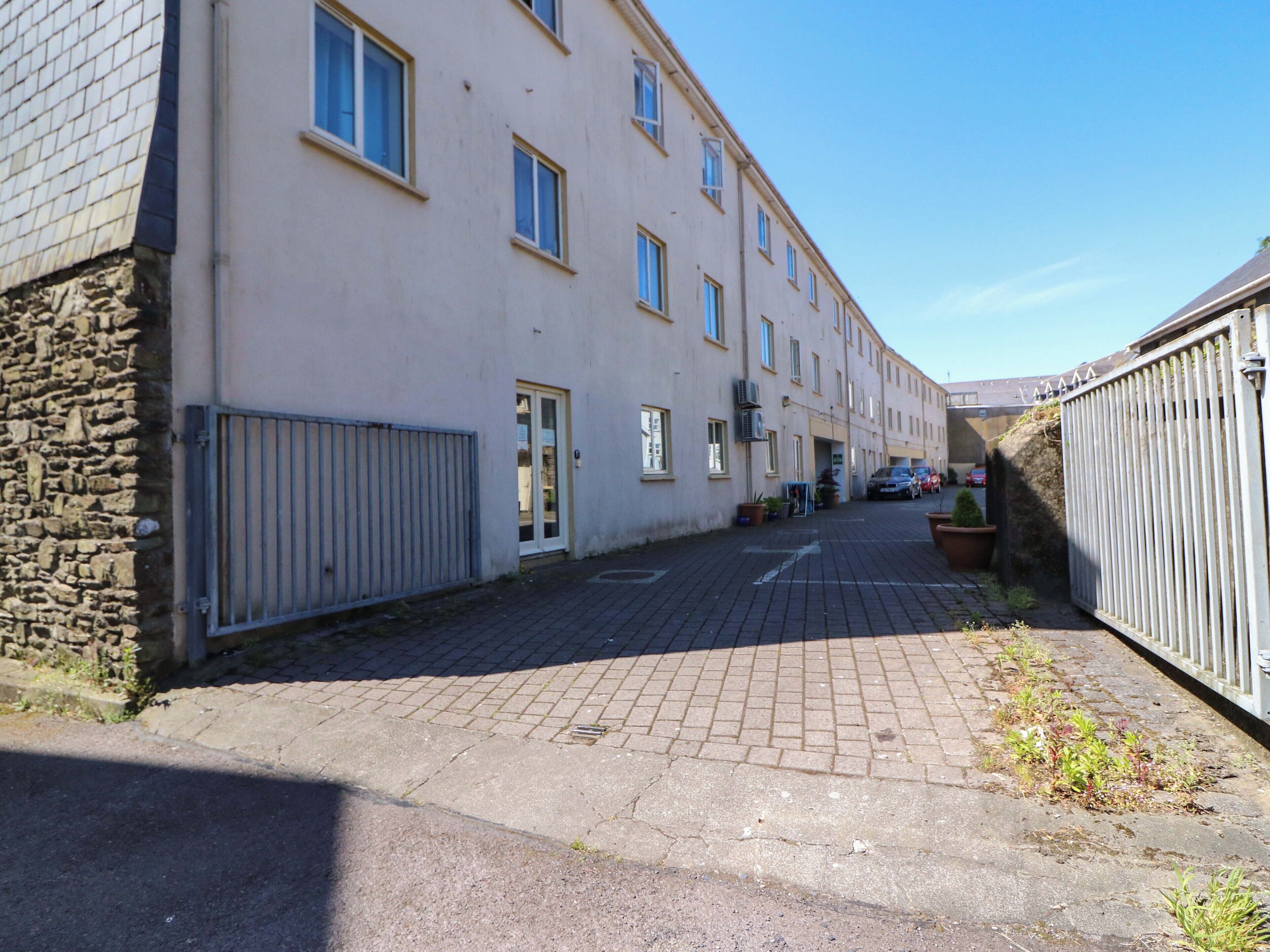 A narrow street with a row of buildings, a stone wall on one side, and a metal fence on the other.
