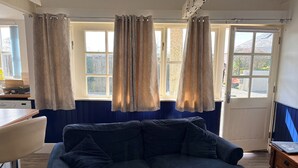 Ein Zimmer mit einem dunkelblauen Sofa, einem Holzespeisetisch mit Stühlen, einer Küchenzeile mit Herd und einem Fenster mit Vorhängen.