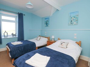 Une chambre avec deux lits simples, une fenêtre avec des rideaux, une petite table avec une lampe et deux tableaux accrochés au mur.